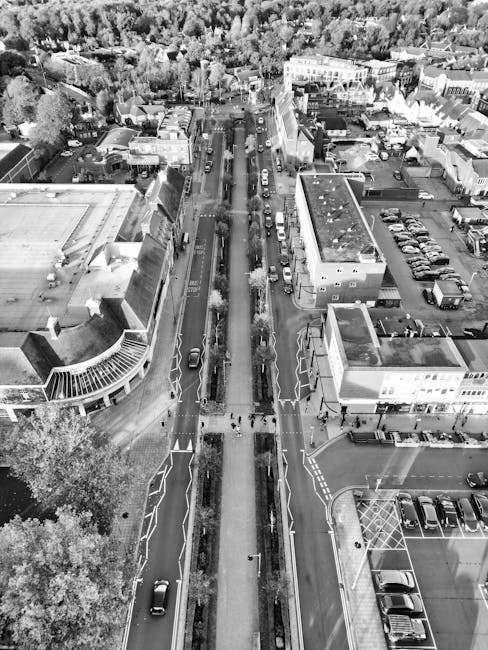 An aerial view of a busy urban street in Uxbridge, showing a wide road with multiple lanes and vehicles parked along the sides, bordered by commercial buildings, retail outlets, and offices. Pedestrians are visible walking on the sidewalks, and a few are crossing the street at designated crossings. The street passes through the town centre, with trees and greenery lining parts of the pavement. On the left side, there is a modern building with large glass windows and an overhanging structure. The right side features additional shops and office spaces, with a parking lot nearby filled with cars. The scene captures the typical environment where house removals and furniture transport might occur during a home relocation or moving service, and Uxbridge Movers could be involved in coordinating the logistics of loading or unloading vehicles in this area.