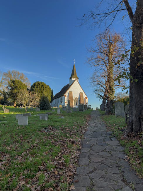 A stone pathway leading up a gentle incline through a churchyard with green grass and fallen leaves, flanked by leafless trees on the right and various headstones scattered across the lawn. In the background, a white church with a pointed steeple and arched windows stands against a clear blue sky, with some darker evergreen trees to the left. The scene is well-lit with natural daylight, emphasizing the peaceful atmosphere of the rural church setting. Uxbridge Movers' house removals team may be involved in transporting furnishings from such locations as part of home relocation or furniture transport services, with their expertise in packing and loading processes evident in the careful handling of items on site.